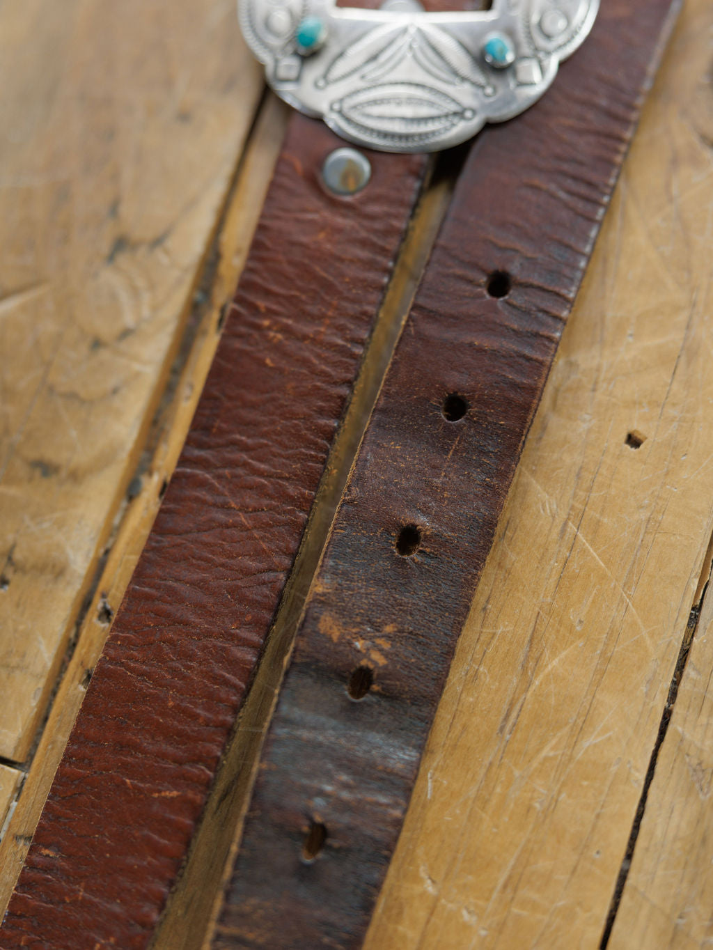 Close-up of the Ruidoso Belt by Savannah Sevens western life{&}style, a worn brown leather belt with multiple holes resting on wood. A partially visible Navajo sterling silver and turquoise buckle is seen at the top.