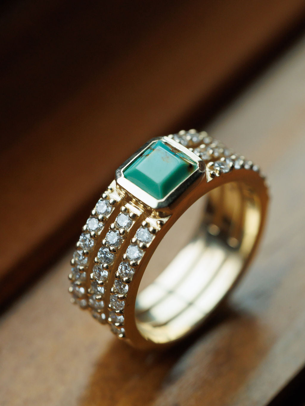 The Savannah Sevens western life{&}style Diamond Turquoise Ring features a vivid turquoise center stone and two rows of sparkling lab-grown diamonds on the band, displayed elegantly on a wooden surface. Collaboration the Devereaux Gold.
