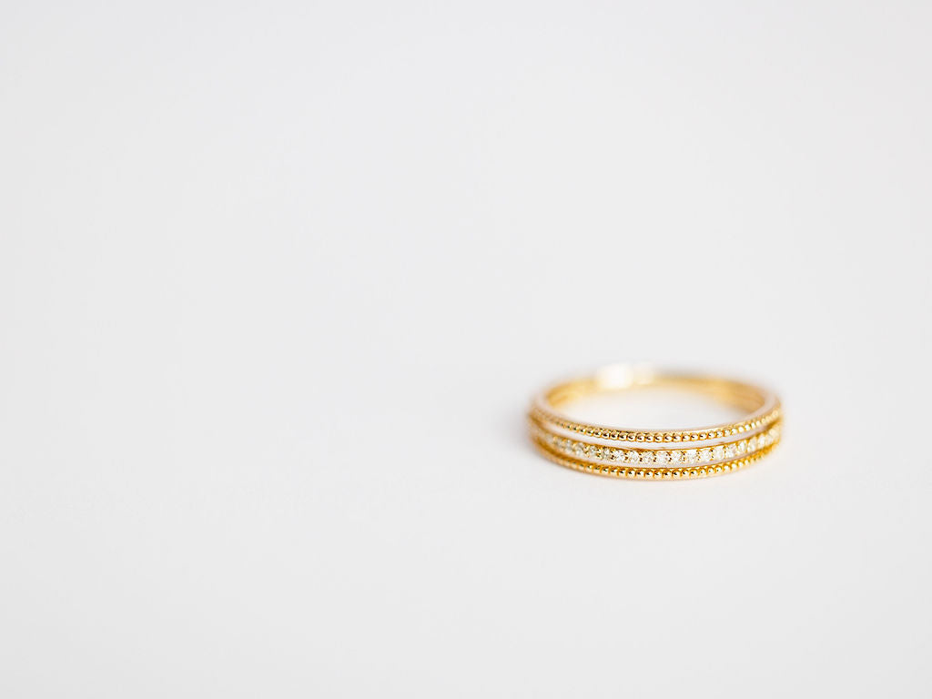 The Diamond Milgrain Ring by Savannah Sevens western life{&}style, crafted in 14K gold with intricate milgrain detailing, sits on a plain white surface, positioned to the right with empty space on the left.