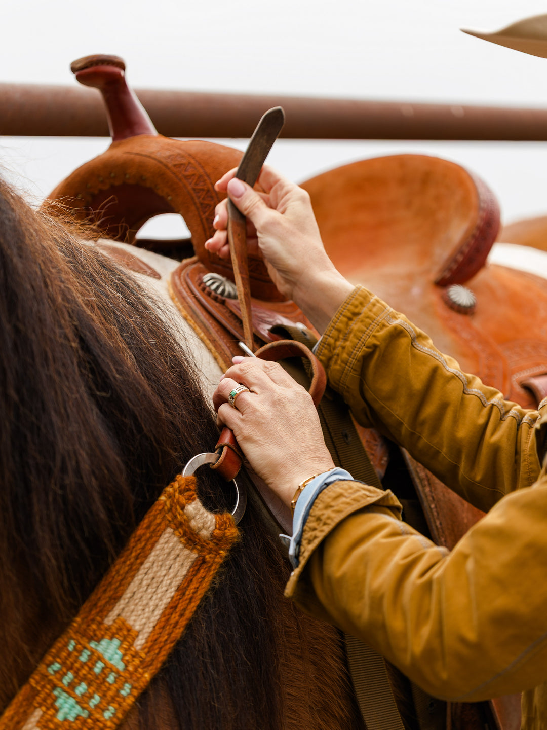 A person in a brown jacket fastens a saddle with decorative stitching on a horse, carefully adjusting the leather tack.
