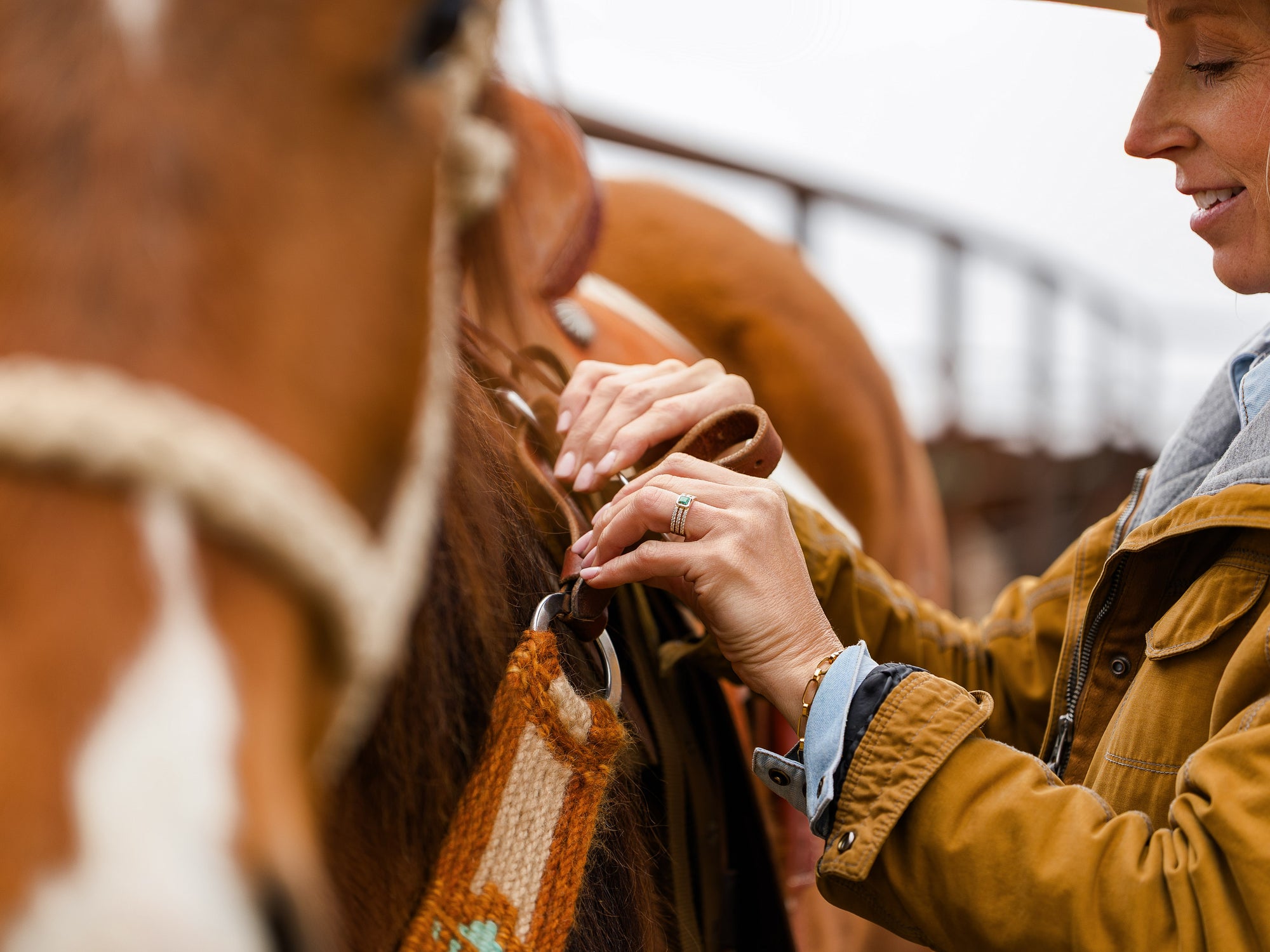 A person in a brown jacket fastens a saddle on a horse outdoors. The scene highlights their hands and the horse’s side, with blurred fencing in the background under natural light.