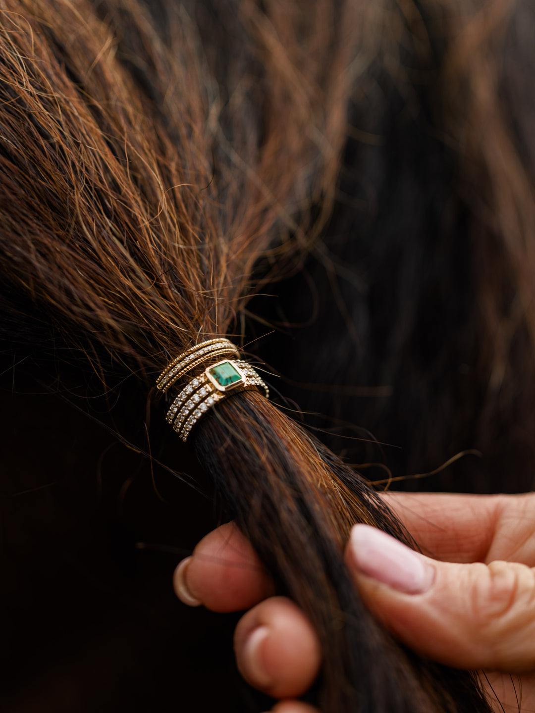 A close-up of a hand holding brown hair, tied with elegant rings, including one adorned with a green gemstone and another encrusted with diamonds, serving as stylish decorative hair accessories.