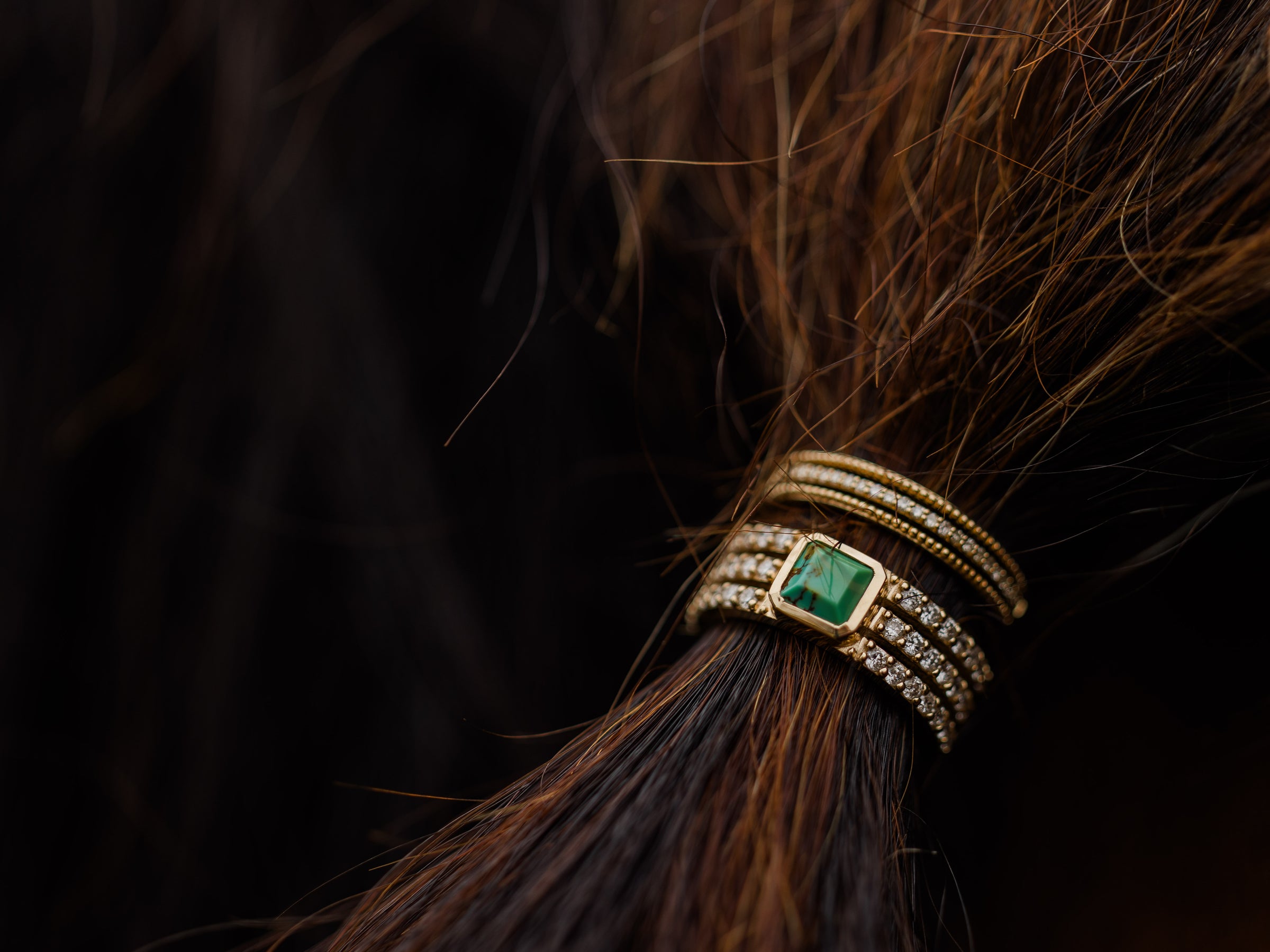 A close-up of brown hair tied with an elegant gold hair accessory featuring multiple bands and a green gemstone, accented by rows of small diamonds.