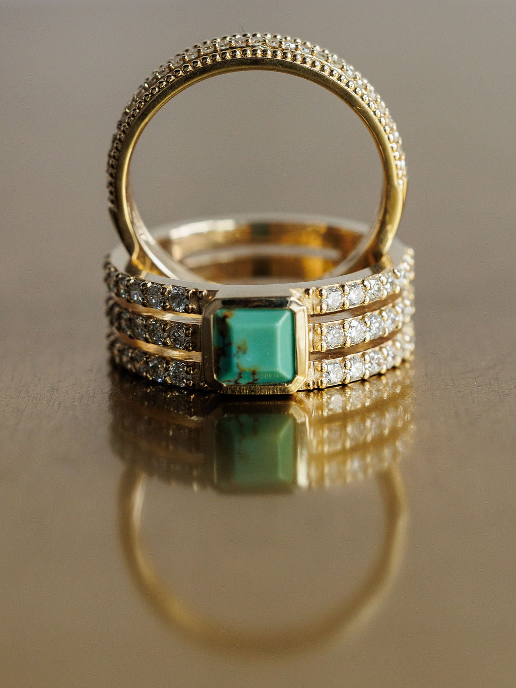 A close-up of Savannah Sevens western life{&}style’s Diamond Turquoise Ring with lab grown diamonds and a rectangular turquoise stone beside a simple beaded-edge band, both displayed on a reflective surface. Collaboration the Devereaux Gold.