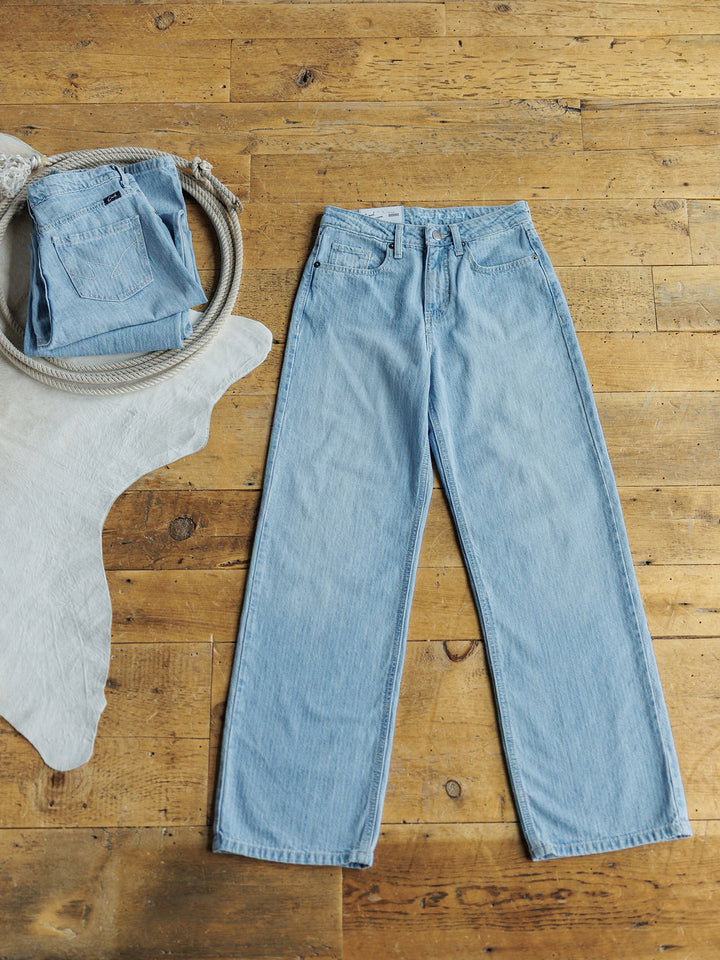 The Landry High Rise Slim Wide Leg Jean by Savannah Sevens western life{&}style in light blue lies flat on a wooden floor beside a white rug. Another pair of pinstripe denim jeans is folded in a woven basket in the top left corner.