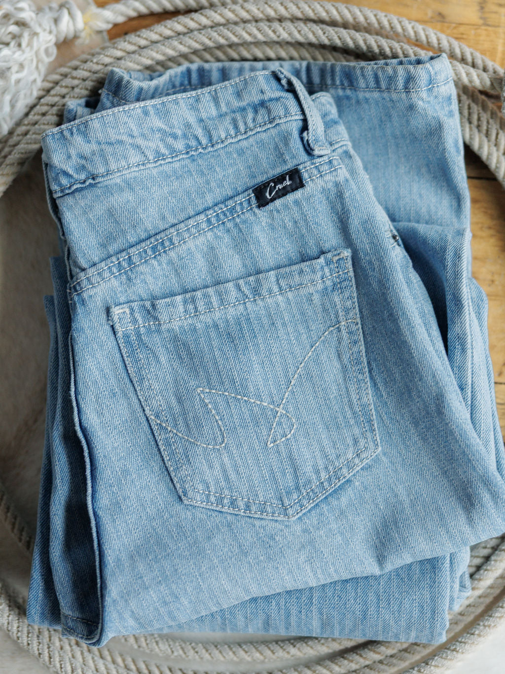 A pair of Savannah Sevens western life{&}style The Landry High Rise Slim Wide Leg Jeans in light blue, neatly folded to show the back pocket and brand label, rests atop coiled rope on a wooden surface.