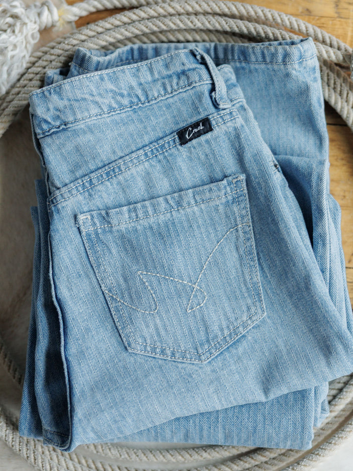 A pair of Savannah Sevens western life{&}style The Landry High Rise Slim Wide Leg Jeans in light blue, neatly folded to show the back pocket and brand label, rests atop coiled rope on a wooden surface.