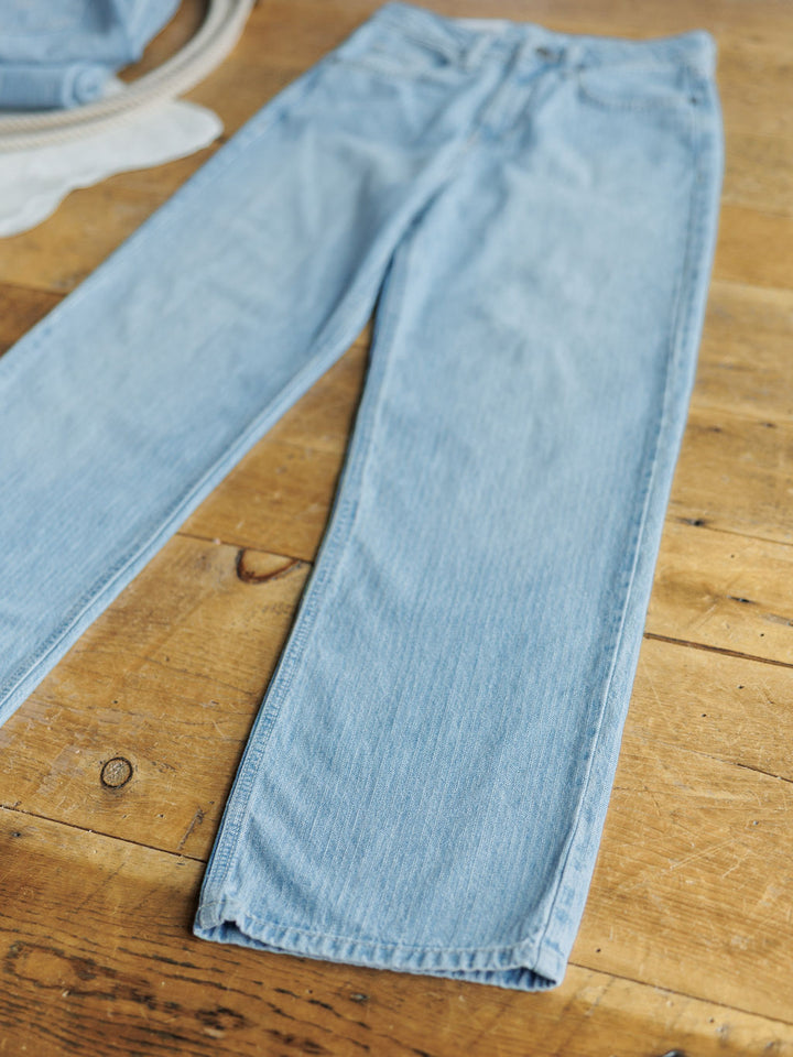 The Landry High Rise Slim Wide Leg Jean by Savannah Sevens western life{&}style is laid flat on a rustic wooden surface, with light-colored fabric visible in the upper left corner.
