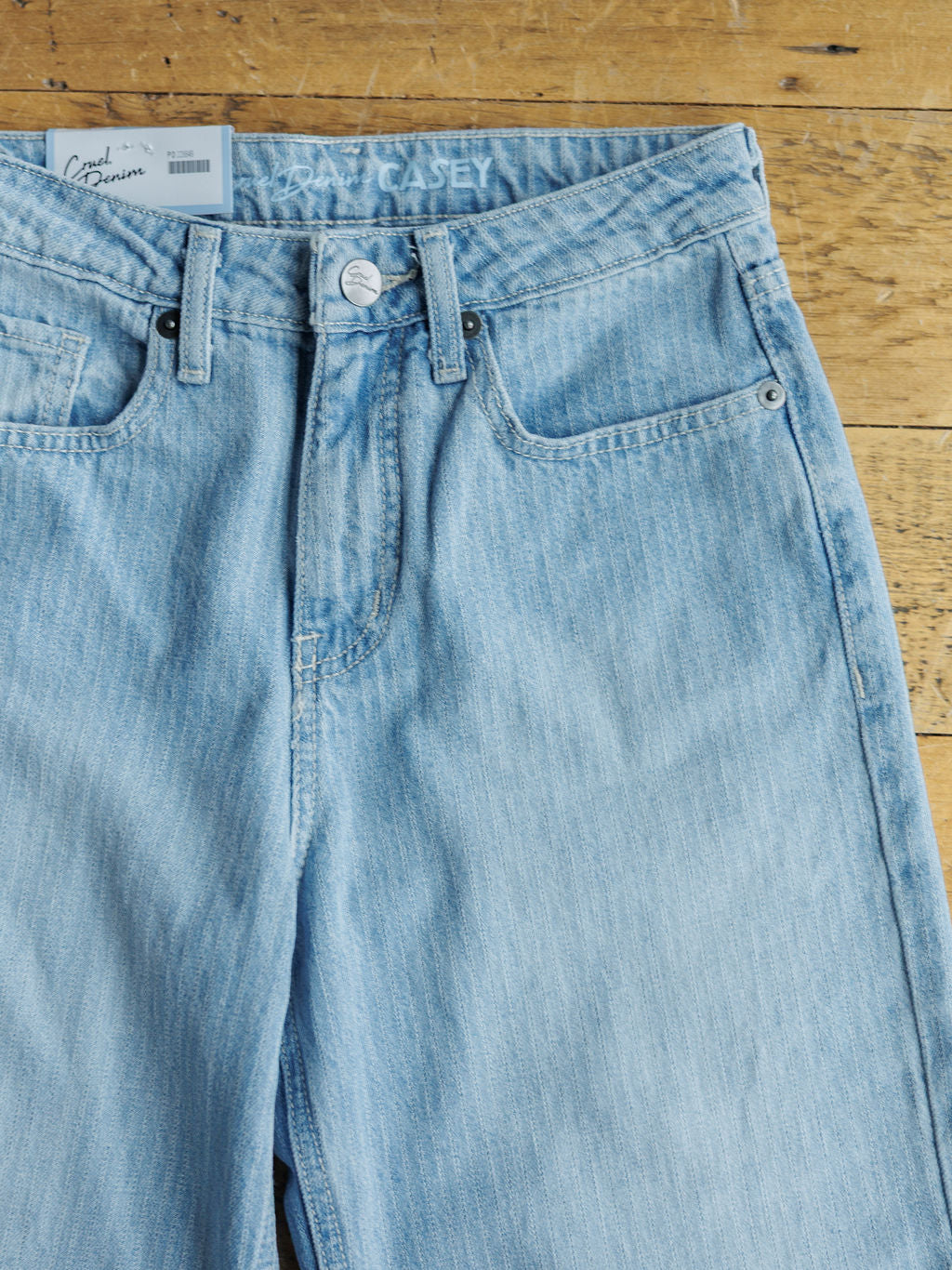 A pair of light blue Savannah Sevens western life{&}style The Landry High Rise Slim Wide Leg Jeans with a button and zipper fly lies flat on wood. A white branded label is on the waistband, and a white price tag is tucked into the left pocket.