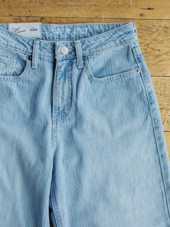 A pair of light blue Savannah Sevens western life{&}style The Landry High Rise Slim Wide Leg Jeans with a button and zipper fly lies flat on wood. A white branded label is on the waistband, and a white price tag is tucked into the left pocket.