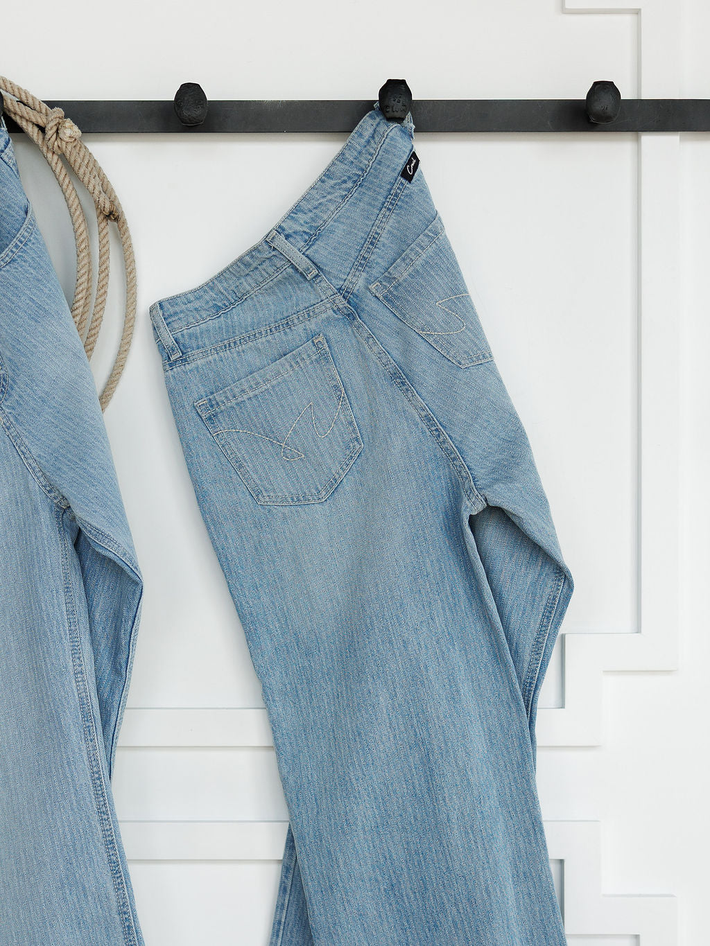 The Landry High Rise Slim Wide Leg Jean in light blue pinstripe by Savannah Sevens western life{&}style hangs on a black hook against a white paneled wall. Another pair of jeans and a coiled rope appear partially on the left.