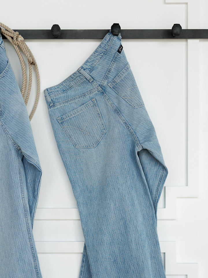 The Landry High Rise Slim Wide Leg Jean in light blue pinstripe by Savannah Sevens western life{&}style hangs on a black hook against a white paneled wall. Another pair of jeans and a coiled rope appear partially on the left.