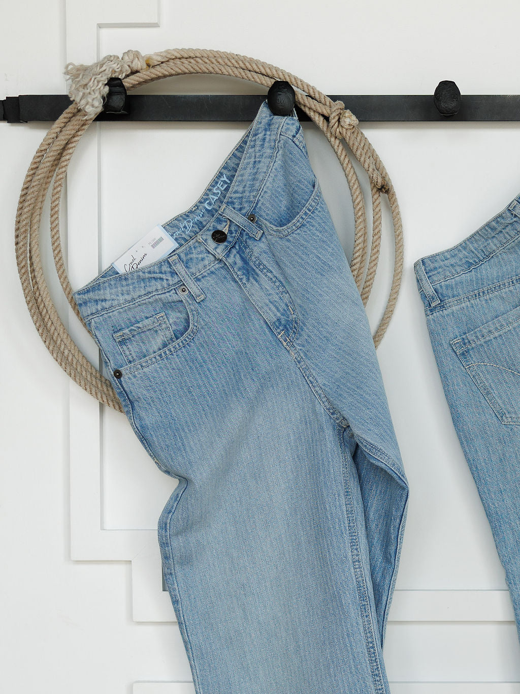 A pair of light blue Savannah Sevens western life{&}style The Landry High Rise Slim Wide Leg Jeans hangs on a black rod with a thick beige rope in the belt loop. Another high rise wide leg jean is partially visible on the right.