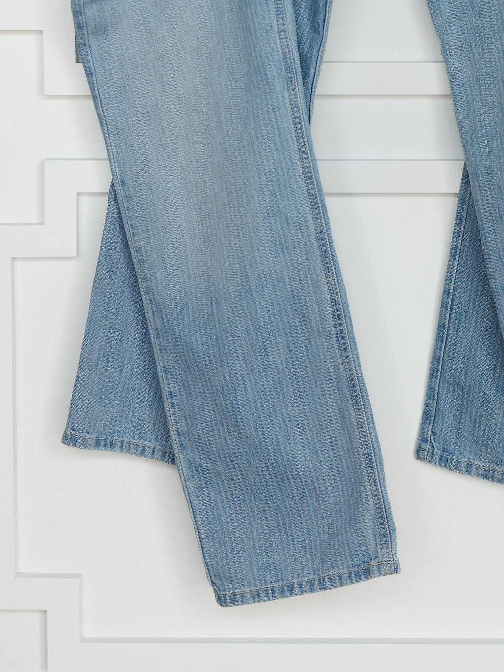 A close-up of The Landry High Rise Slim Wide Leg Jean by Savannah Sevens western life{&}style, showing the lower legs in light blue pinstripe denim with one pant leg overlapping the other against a white panel wall.