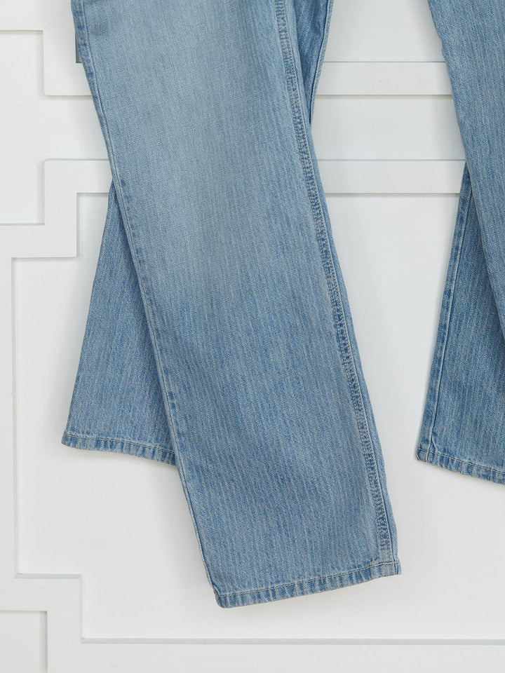 A close-up of The Landry High Rise Slim Wide Leg Jean by Savannah Sevens western life{&}style, showing the lower legs in light blue pinstripe denim with one pant leg overlapping the other against a white panel wall.