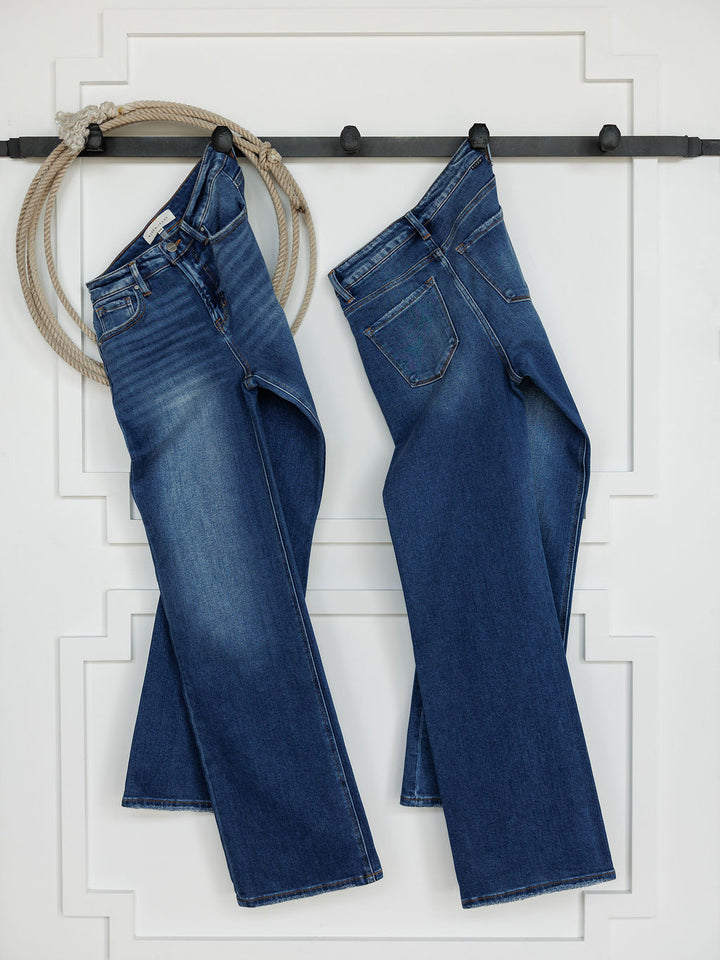 Two pairs of The Bristol High Rise 90's Wide Leg Jeans by Savannah Sevens western life{&}style hang by their belt loops from a black rod on a white wall, with a coiled rope on the left. They feature an ultra high rise and subtle thigh fading.