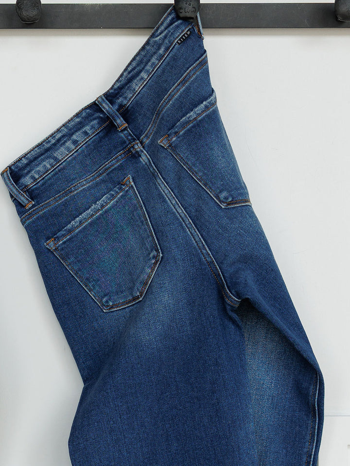 The Bristol High Rise 90's Wide Leg Jean by Savannah Sevens western life{&}style hangs by the belt loop on a black metal hook, displaying its blue wash, wide leg fit, ultra high rise waistband, and visible back pockets against a white background.