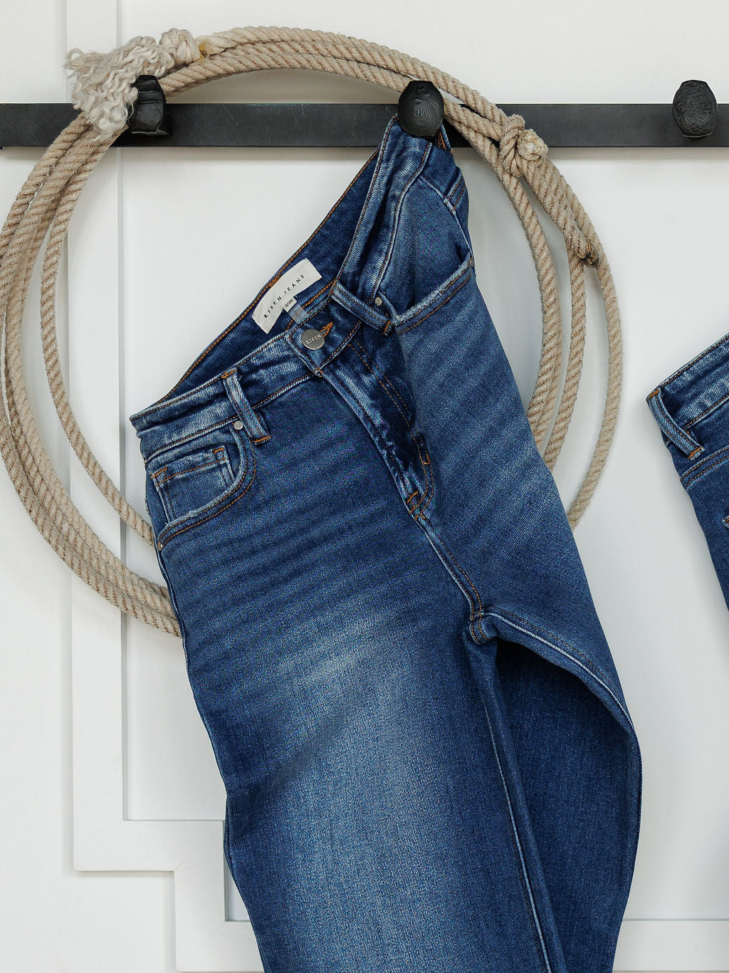 A pair of The Bristol High Rise 90's Wide Leg Jeans by Savannah Sevens western life{&}style hangs on a black wall hook, with a coiled rope looped around the hook in the background.