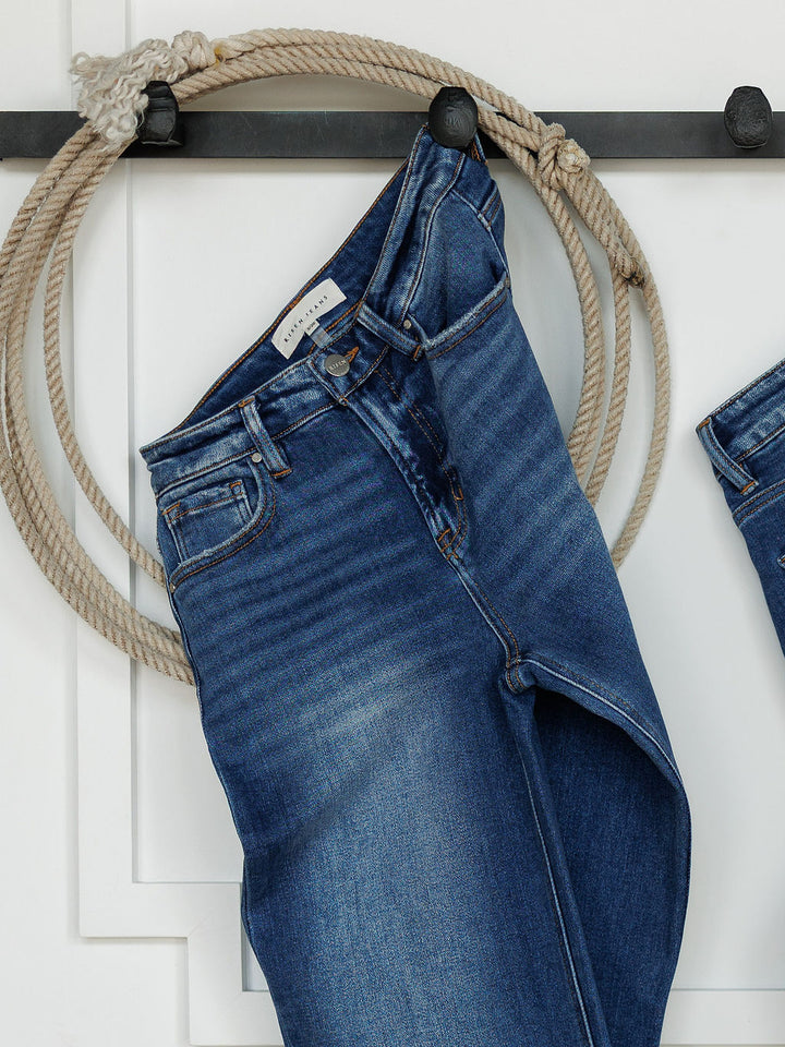 A pair of The Bristol High Rise 90's Wide Leg Jeans by Savannah Sevens western life{&}style hangs on a black wall hook, with a coiled rope looped around the hook in the background.