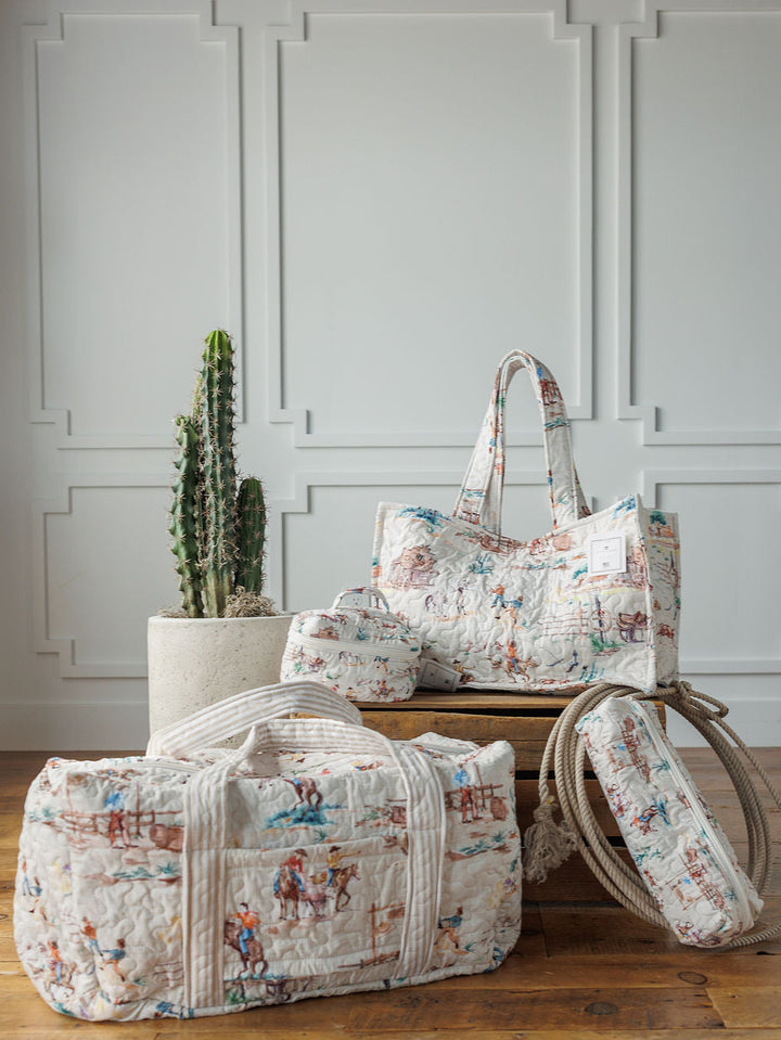 The Retro Cowboy Collection by Savannah Sevens western life&style: a handmade quilted bag set with vintage prints—duffel, tote, cosmetic case, and cylinder bag—displayed on a wooden bench by a cactus in a bright, paneled room.