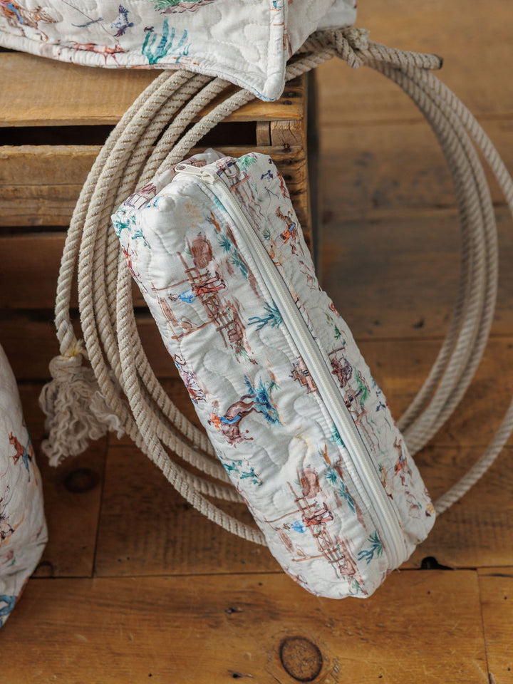 The Retro Cowboy Collection by Savannah Sevens western life&style—a quilted bag set with a white zipper and colorful cowboy and horse print—rests on coiled rope atop a wooden crate on a wooden floor.