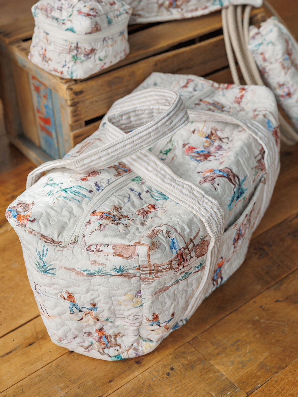 The Retro Cowboy Collection by Savannah Sevens western life&style showcases a handmade quilted duffel bag with a cowboy and horse print, displayed on a wooden floor beside rustic crates and coordinating accessories.