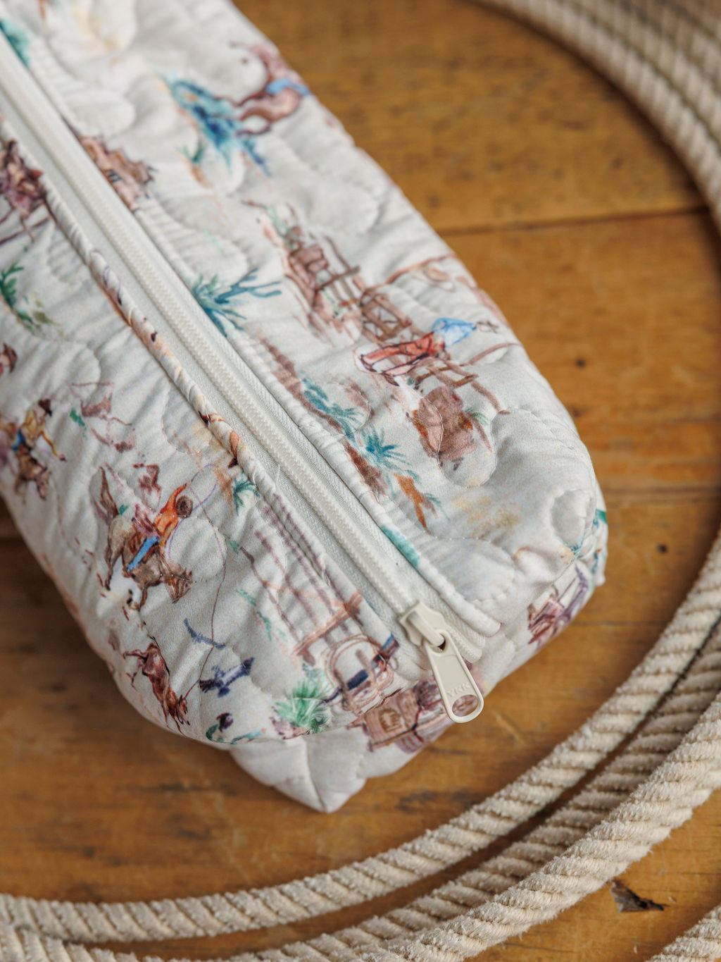 A handmade zippered pouch from the Retro Cowboy Collection by Savannah Sevens western life&style, featuring a colorful scenic print, shown on a wooden surface with a coiled rope.
