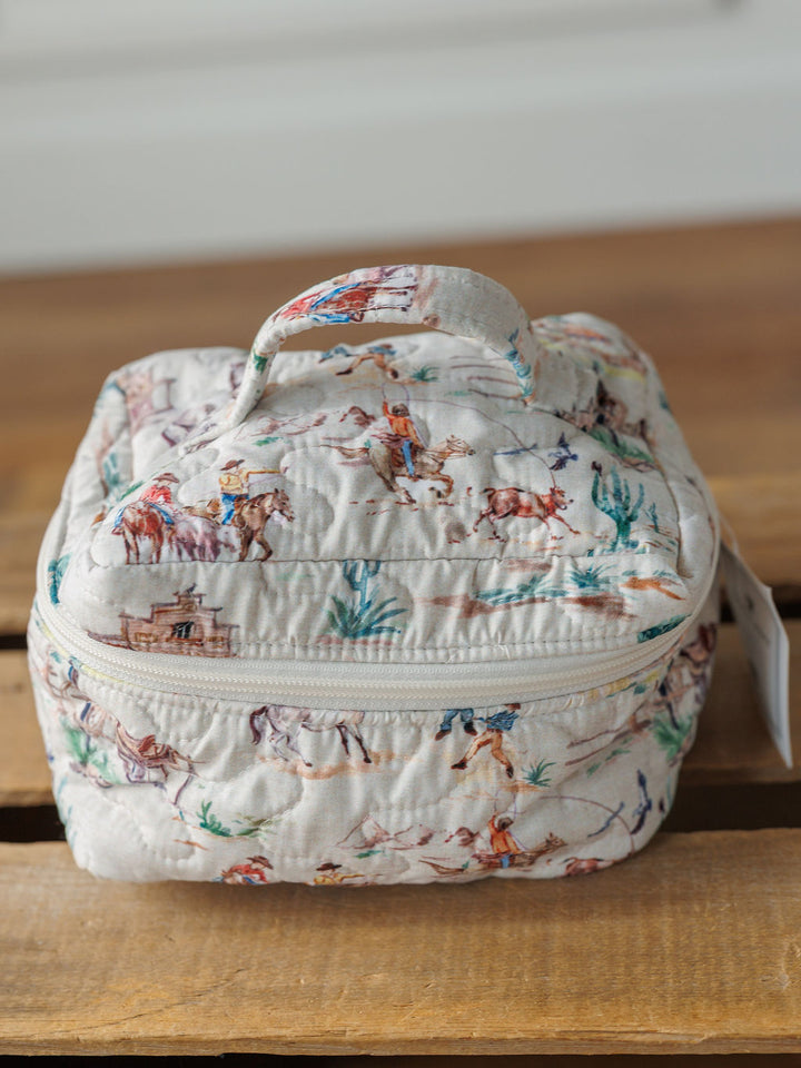 A small quilted fabric bag from the Retro Cowboy Collection by Savannah Sevens western life&style, featuring cowboys, horses, and desert scenery, with a zipper and handle, sits on a wooden surface.