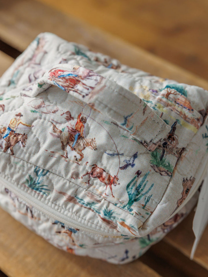 The Retro Cowboy Collection bag from Savannah Sevens western life{&}style features quilted fabric, a handle, and a vibrant cowboy scene print, shown resting on a wooden surface.