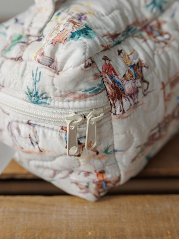 Close-up of a handmade quilted bag from the Retro Cowboy Collection by Savannah Sevens western life&style, with a double zipper and colorful cowboys and horses print on cream, resting on a wooden surface.