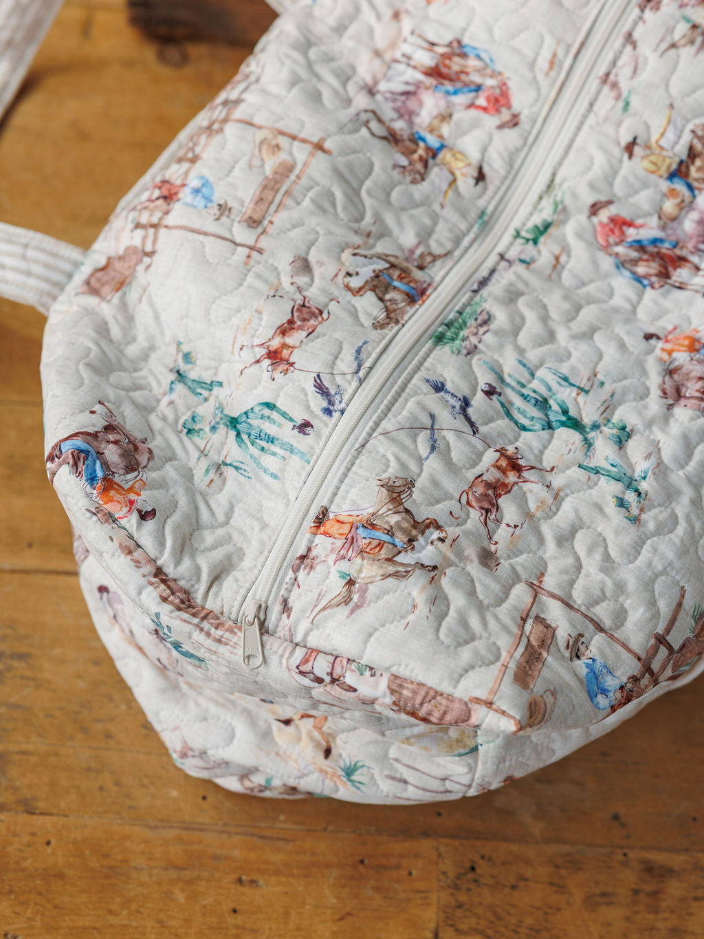 A close-up of a handmade quilted bag set from the Retro Cowboy Collection by Savannah Sevens western life&style, featuring colorful cowboy and horse illustrations on a light background, with a white zipper and displayed on wood.