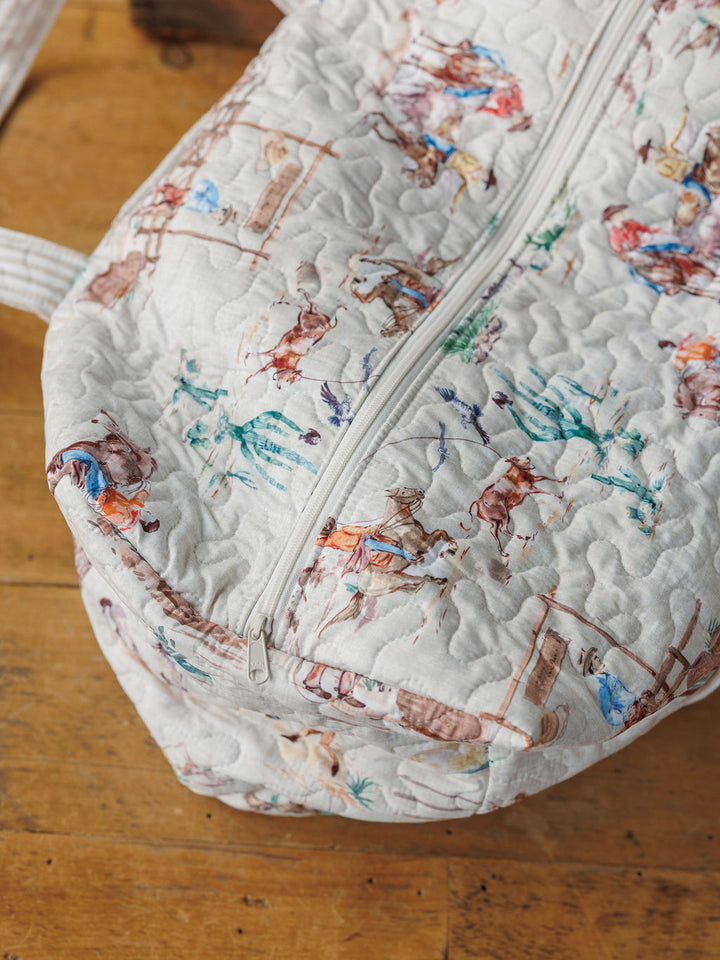 A close-up of a handmade quilted bag set from the Retro Cowboy Collection by Savannah Sevens western life&style, featuring colorful cowboy and horse illustrations on a light background, with a white zipper and displayed on wood.