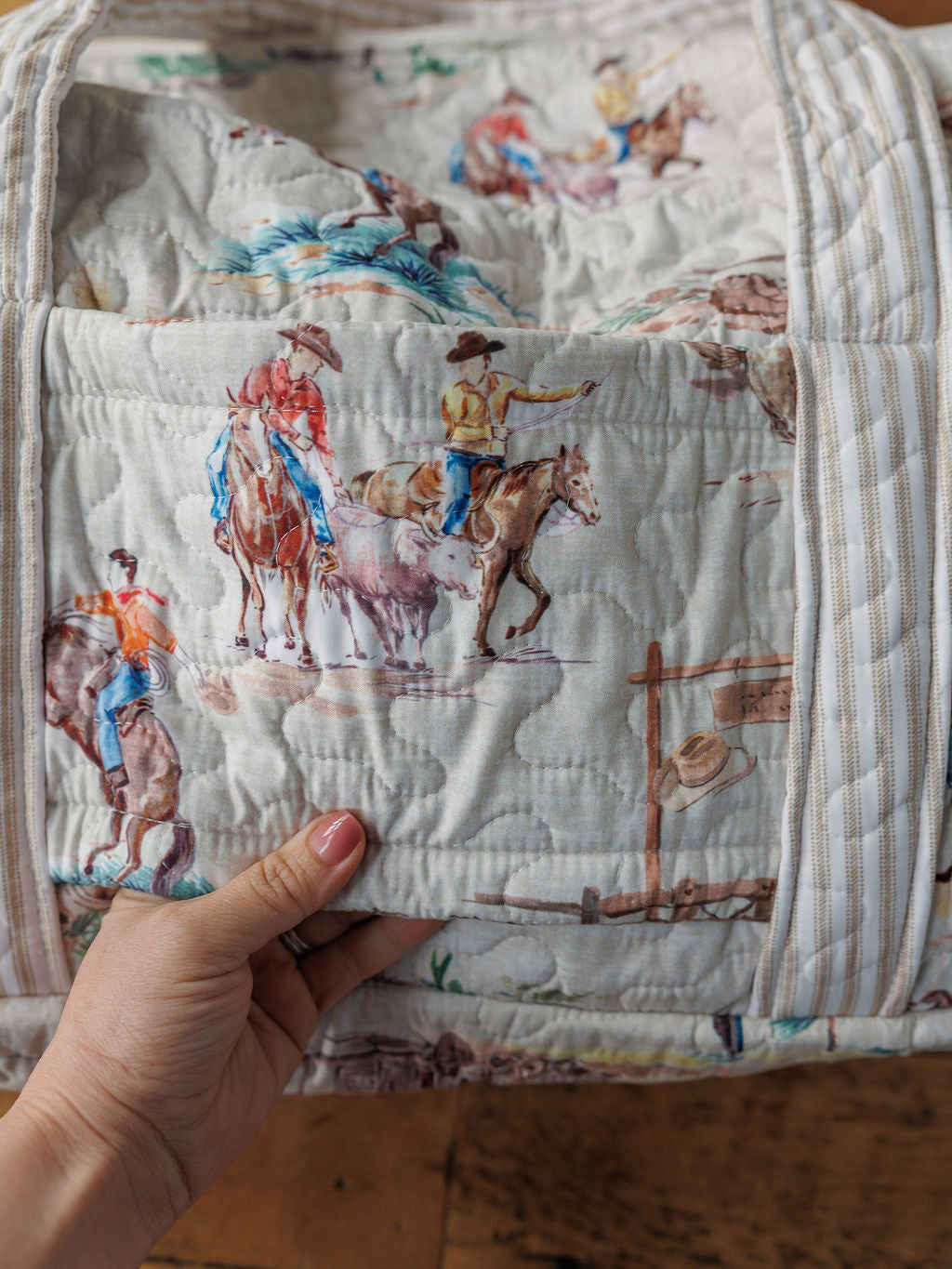 A hand holds a handmade quilt from the Retro Cowboy Collection by Savannah Sevens western life & style, featuring cowboy and cattle illustrations on a light background—a charming piece of western décor.