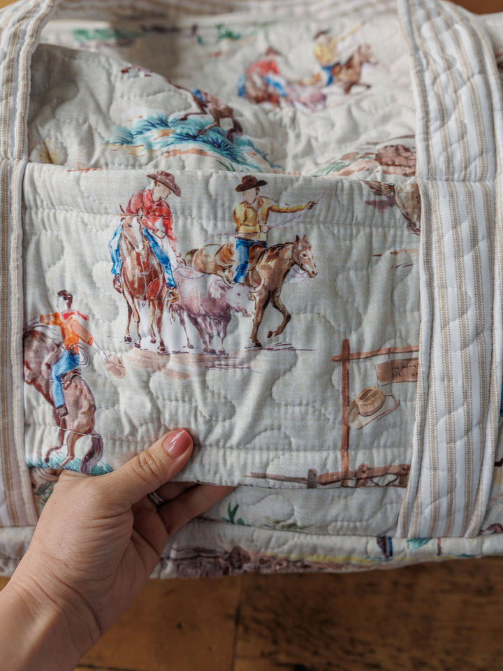 A hand holds a handmade quilt from the Retro Cowboy Collection by Savannah Sevens western life & style, featuring cowboy and cattle illustrations on a light background—a charming piece of western décor.