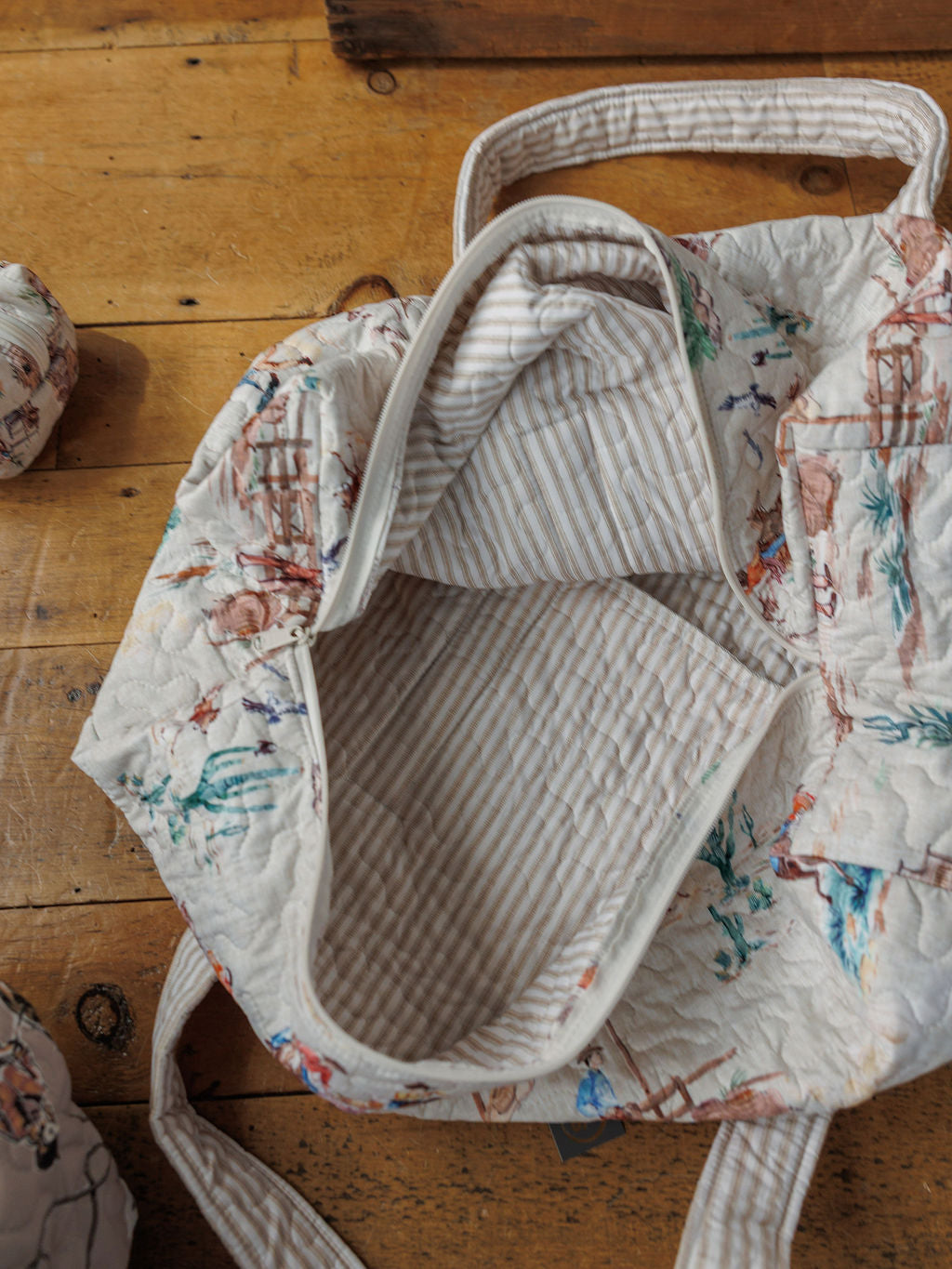 A handmade quilt tote bag from the Retro Cowboy Collection by Savannah Sevens western life&style sits open on a wooden floor, revealing its striped lining. The bag features two handles and a zipper opening with a cream and pastel pattern.