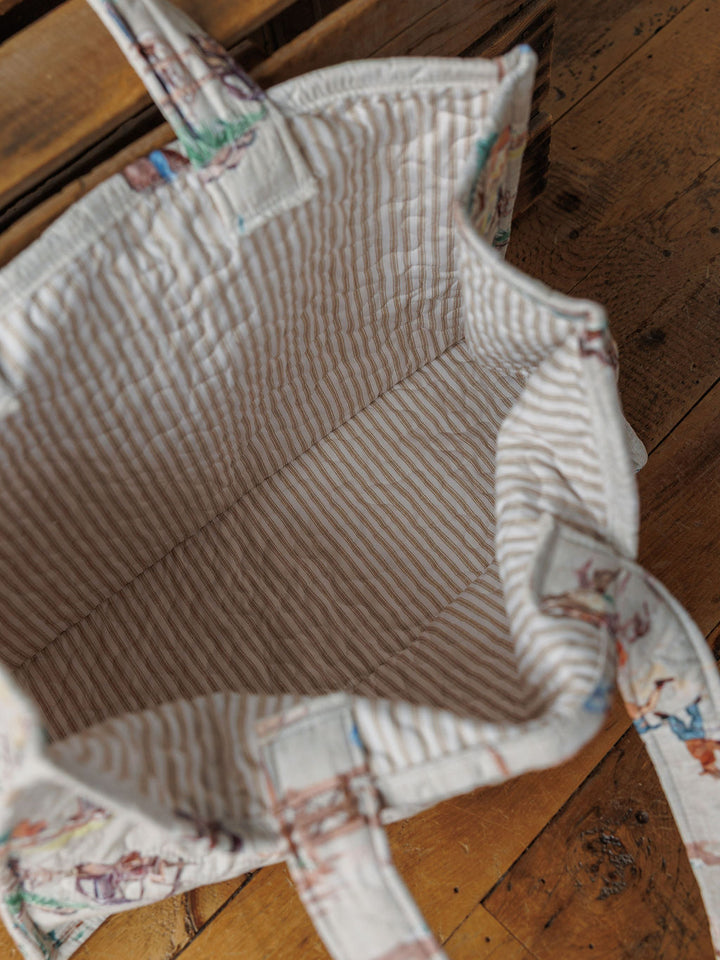 The Retro Cowboy tote by Savannah Sevens western life&style is open to show its spacious, striped-lined interior. Featuring a handle, the handmade quilted bag sits on a wooden floor near furniture from the same collection.