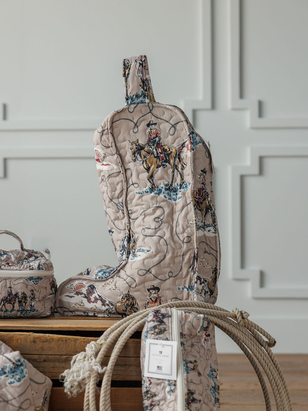 The Montana Summer Collection by Savannah Sevens western life&style features a handmade quilted bag set with cowboy and horse illustrations, displayed upright with matching accessories and a coiled rope against a white paneled wall.