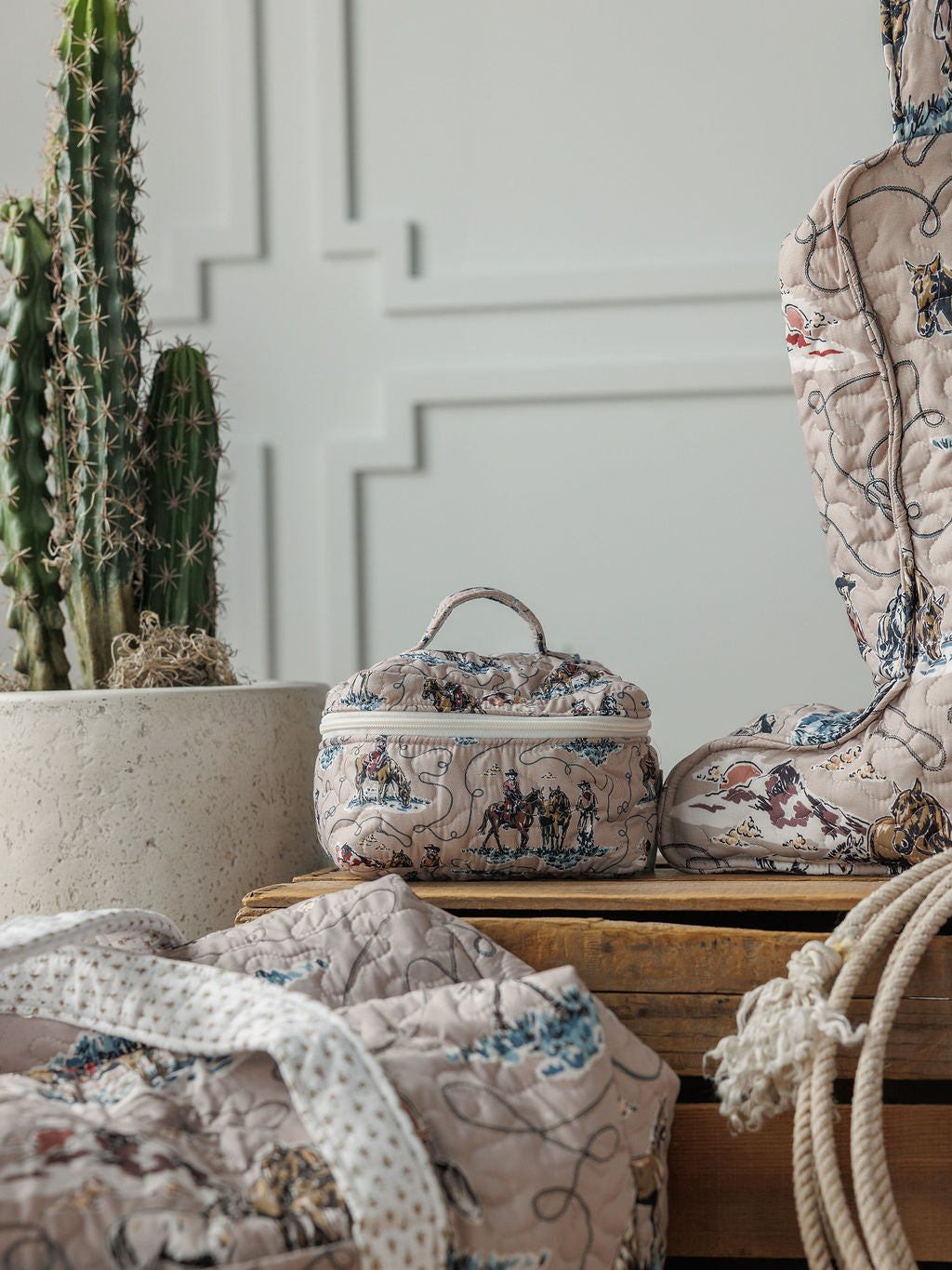 Soft quilted bags and a small box with whimsical prints from the Montana Summer Collection by Savannah Sevens western life&style are arranged on wood beside a tall cactus, set against a textured white wall.