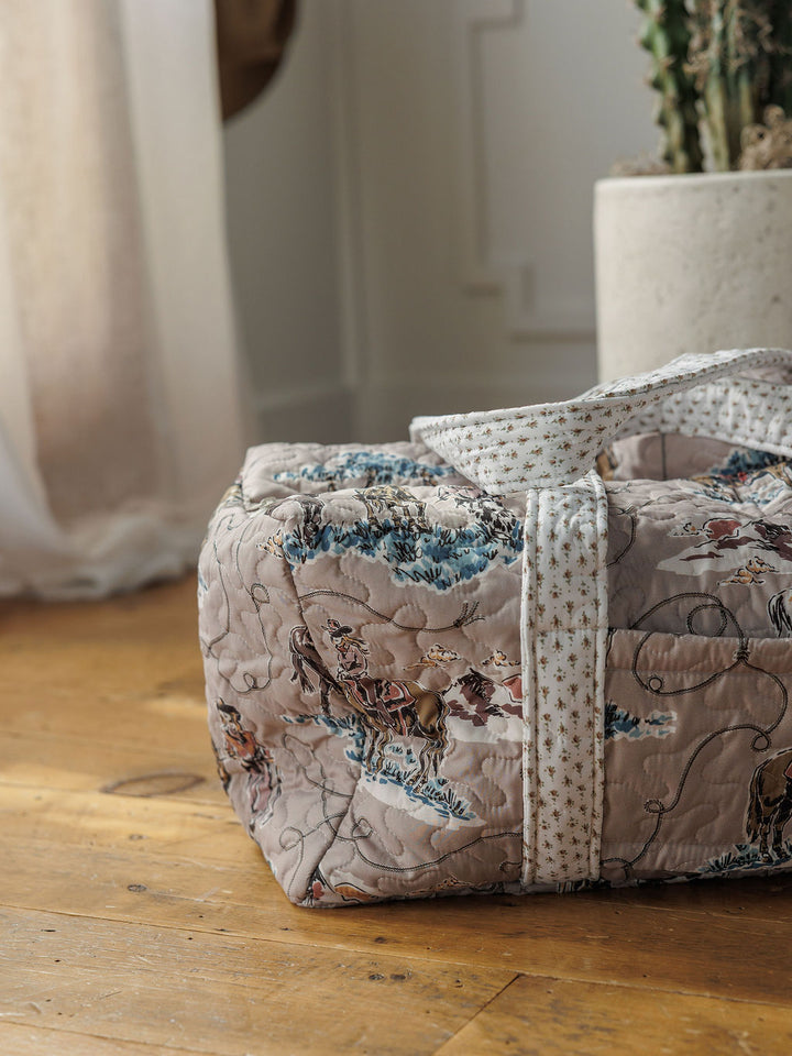 A quilted bag set with a toile pattern and star-patterned handles from the Montana Summer Collection by Savannah Sevens western life&style rests on a wooden floor near a white curtain and large potted cactus.