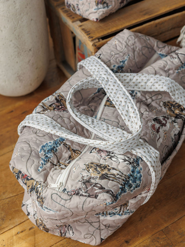 The Montana Summer Collection by Savannah Sevens western life&style includes a light gray quilted duffel with blue and brown floral prints, white zipper, patterned straps, and a matching small bag—both styled on wood surfaces.