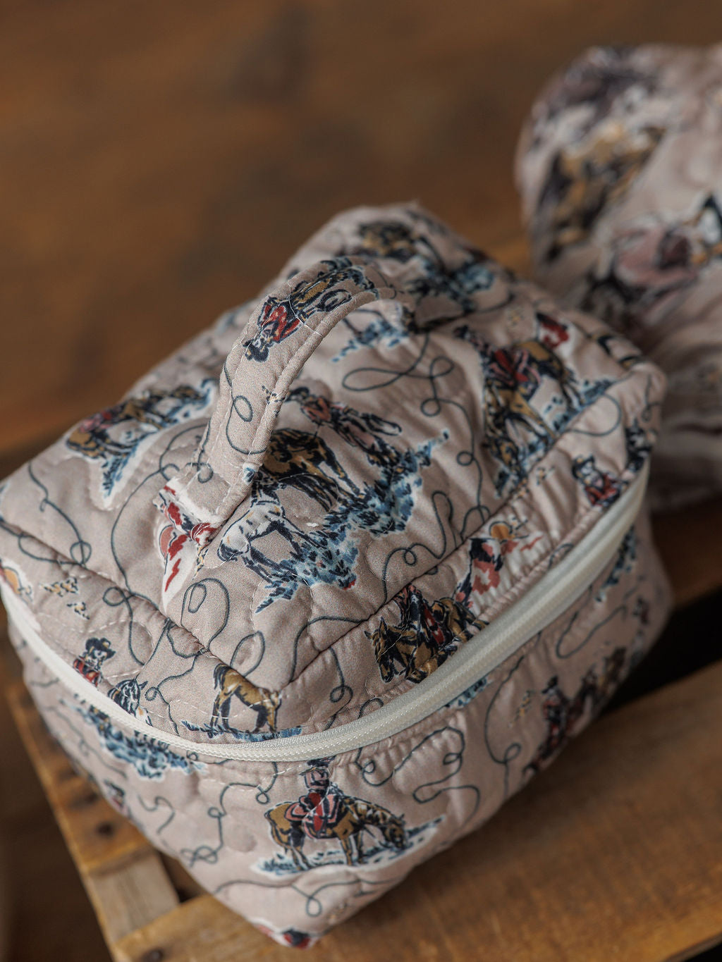 The Montana Summer Collection by Savannah Sevens western life&style is a quilted bag set with zippered closure, featuring cowboy and horse prints—ideal for your western travel collection. Displayed on a wooden surface.