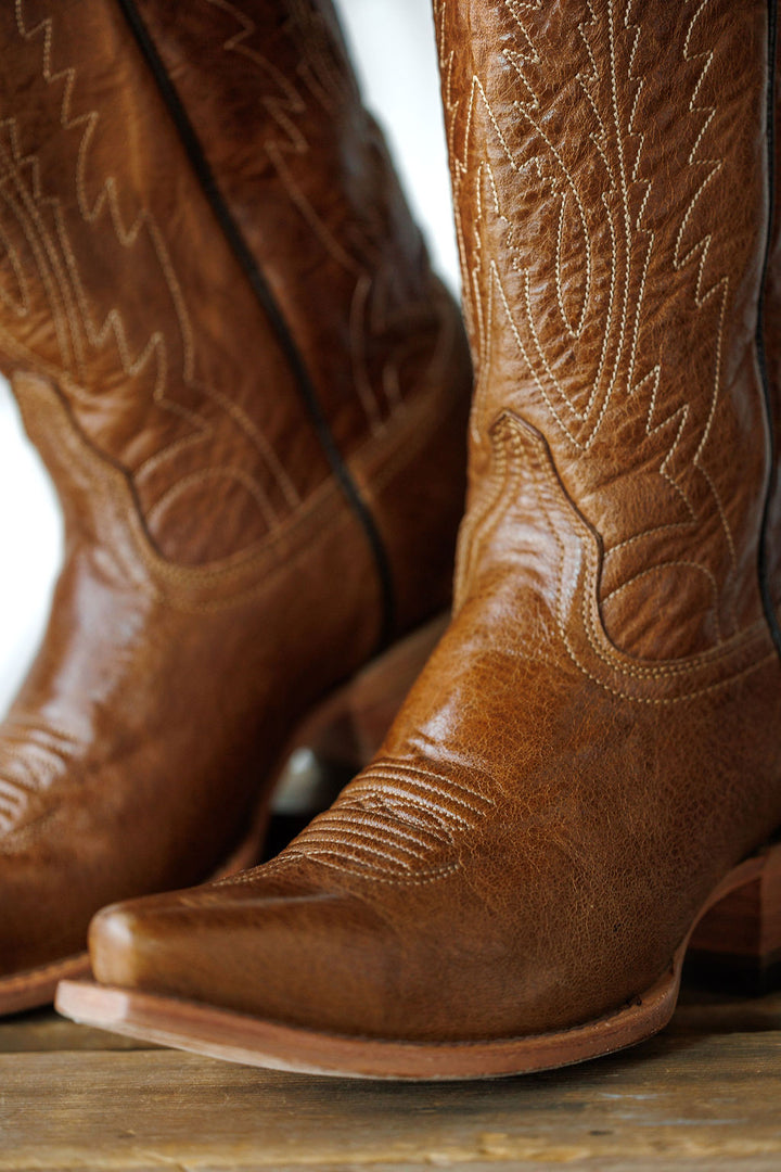 Close-up of The Balcones boots by Savannah Sevens western life{&}style, crafted from pecan leather with decorative stitching, pointed toes, and slightly angled heels, resting on a wooden surface.