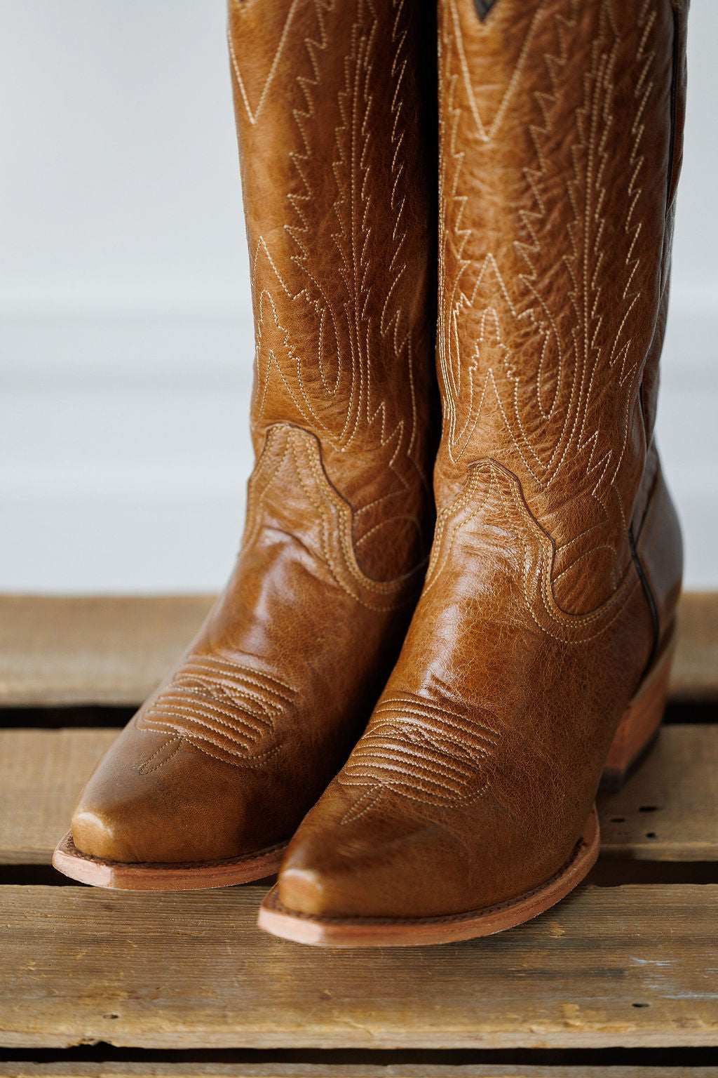 A close-up of The Balcones western boots by Savannah Sevens western life{&}style, crafted from pecan leather with decorative stitching, resting on a wooden surface.