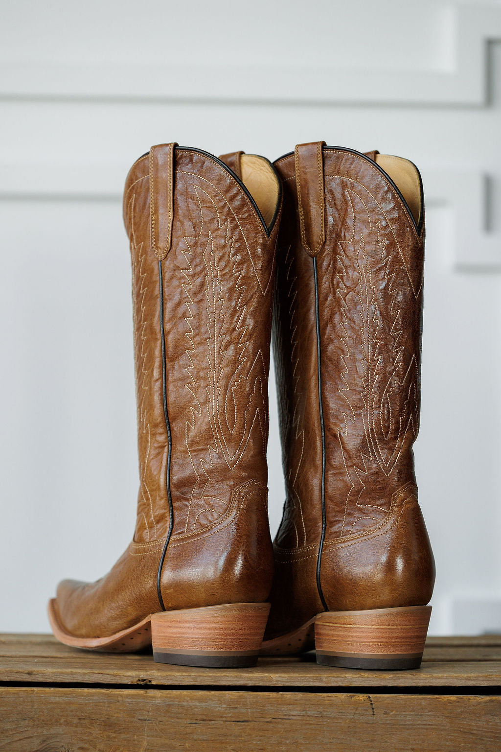 The Balcones boots by Savannah Sevens western life{&}style—tall brown western boots with decorative stitching—stand side by side on a wooden surface, viewed from the back against a light background with white trim.
