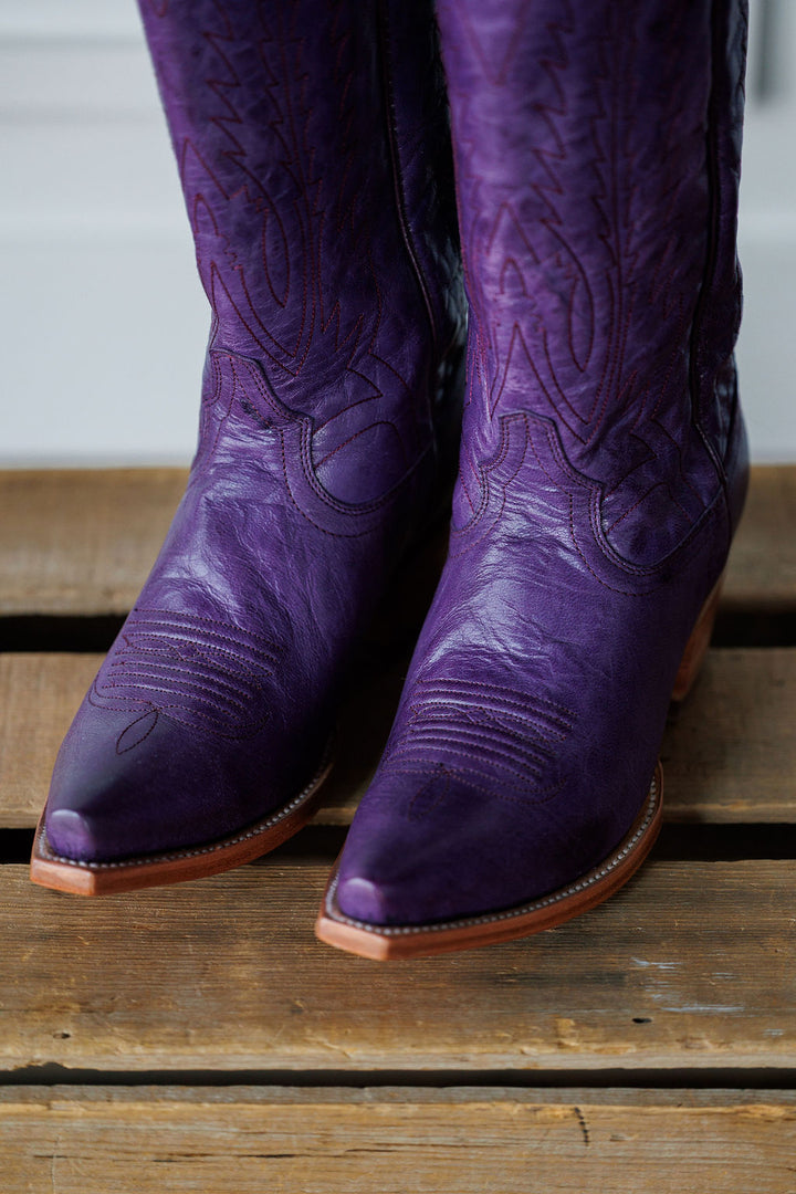 A close-up of The Maricopa plum leather snip toe western boots by Savannah Sevens western life{&}style, featuring decorative stitching, resting on a wooden surface. Made by Black Star. 