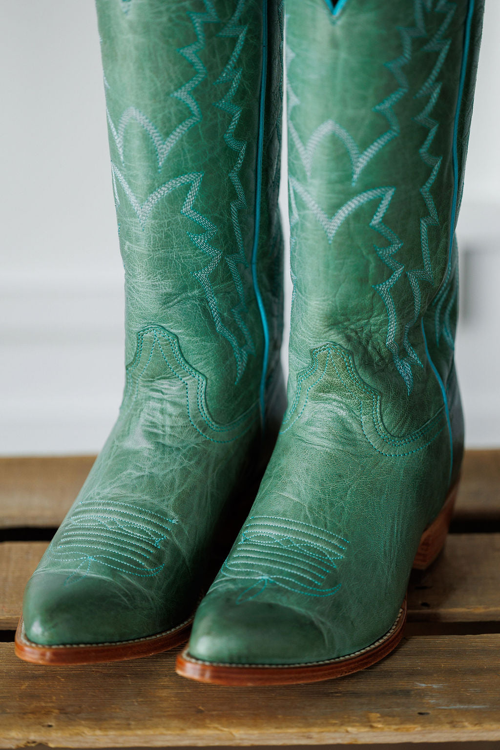 A close-up of The Carico by Savannah Sevens western life{&}style: a green turquoise leather western boot with blue stitching, pointed toes, a tall patterned shaft, shown on a wooden surface.