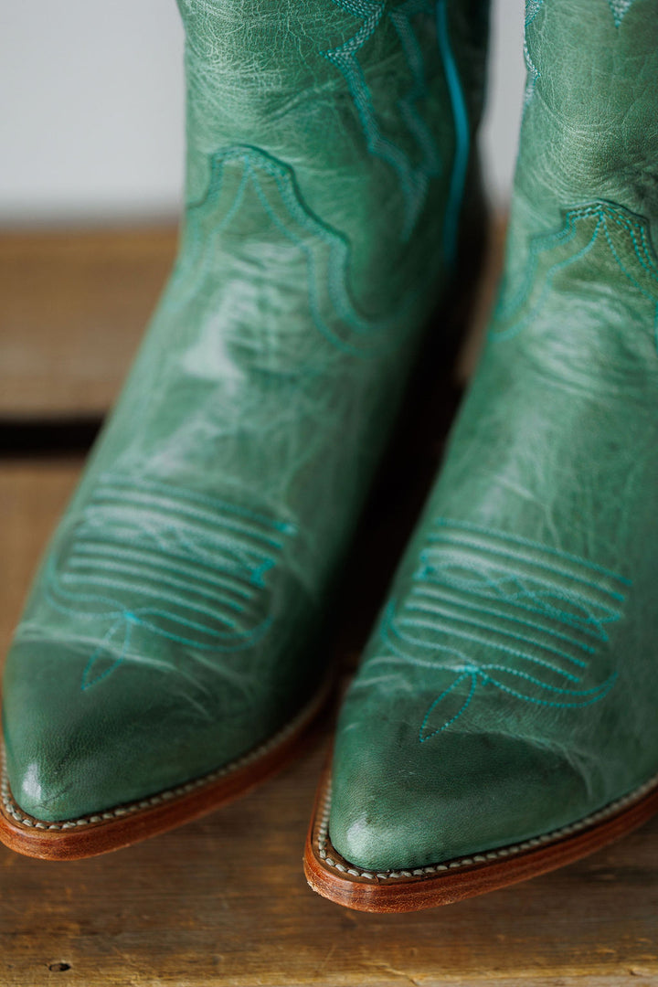 Close-up of the pointed toes of The Carico green turquoise leather western boot by Savannah Sevens western life{&}style, featuring light blue stitching, resting on a wooden surface.