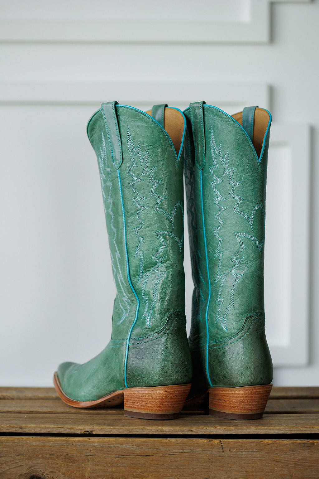 The Carico by Savannah Sevens western life{&}style: tall green turquoise leather western boots with decorative stitching and wooden heels, displayed on a wooden surface against a white paneled background.