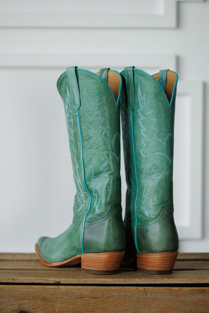 The Carico by Savannah Sevens western life{&}style: tall green turquoise leather western boots with decorative stitching and wooden heels, displayed on a wooden surface against a white paneled background.