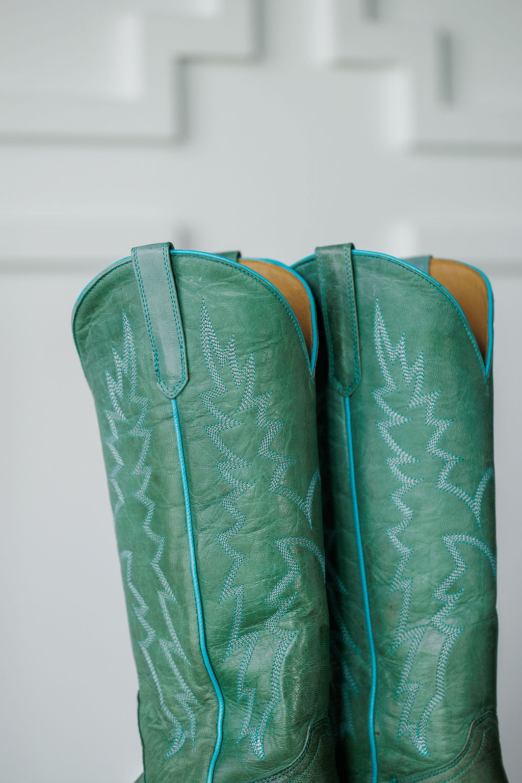 Close-up of The Carico, tall green turquoise leather western boots by Savannah Sevens western life{&}style, featuring light blue decorative stitching and piping, set against a white background with a geometric pattern.